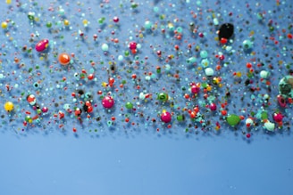 A collection of multicolored paint splatters on a smooth, light blue surface, featuring a variety of shapes and sizes. The colors include vibrant reds, greens, pinks, blues, and oranges, creating a visually stimulating and playful arrangement.