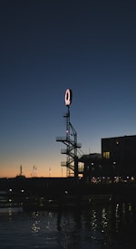 A tall metal sign with a large red letter 'Q' is prominently displayed against a darkening sky, positioned next to a body of water. The sky transitions from deep blue at the top to a warm orange near the horizon, typical of dusk. The silhouette of nearby buildings and a distant docked ship can be seen.
