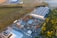An aerial view of an industrial area, featuring large warehouses surrounded by greenery. Various construction vehicles and equipment are neatly arranged in the open yard. The landscape shows a blend of industrial and natural elements, with trees and green spaces adjacent to the structures.
