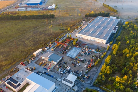 An aerial view of an industrial area, featuring large warehouses surrounded by greenery. Various construction vehicles and equipment are neatly arranged in the open yard. The landscape shows a blend of industrial and natural elements, with trees and green spaces adjacent to the structures.