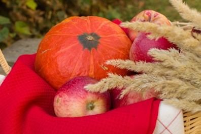 A wicker basket contains a large orange pumpkin, several red apples, and stalks of wheat or dried grass. The basket is lined with a red cloth, adding a vibrant contrast to the autumnal colors of the fruits and the natural elements.