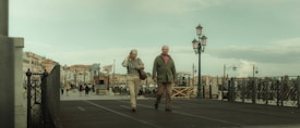 An elderly couple walks leisurely on a boardwalk, surrounded by street lamps and overlooking a waterfront area with buildings in the background. The sky is overcast, giving the scene a muted, tranquil atmosphere.