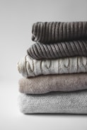 A neatly folded stack of cozy, stylish sweaters in neutral tones on a wooden table.