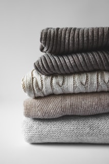 A stack of folded sweaters arranged vertically, showcasing different knitting textures and patterns. The sweaters display a range of neutral colors, including gray and beige tones.