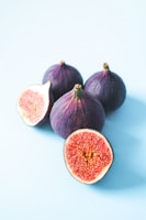 Four whole figs with deep purple skin and a cut fig displaying a rich, pinkish-red interior are arranged on a light blue background. The figs are slightly glossy and textured, with one cut in half to show the intricate pattern of seeds inside.