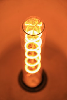 A close-up view of an illuminated incandescent light bulb with a visible spiral filament radiating a warm, orange glow. The bulb is encased in a cylindrical glass tube with a slight blurring towards the bottom, and the background is uniformly warm-toned.