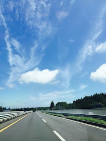 A clear highway stretches into the distance under a vast blue sky with a few scattered clouds. The road is flanked by metal guardrails and greenery, with trees lining both sides. The perspective suggests motion and depth, giving a sense of travel and open space.