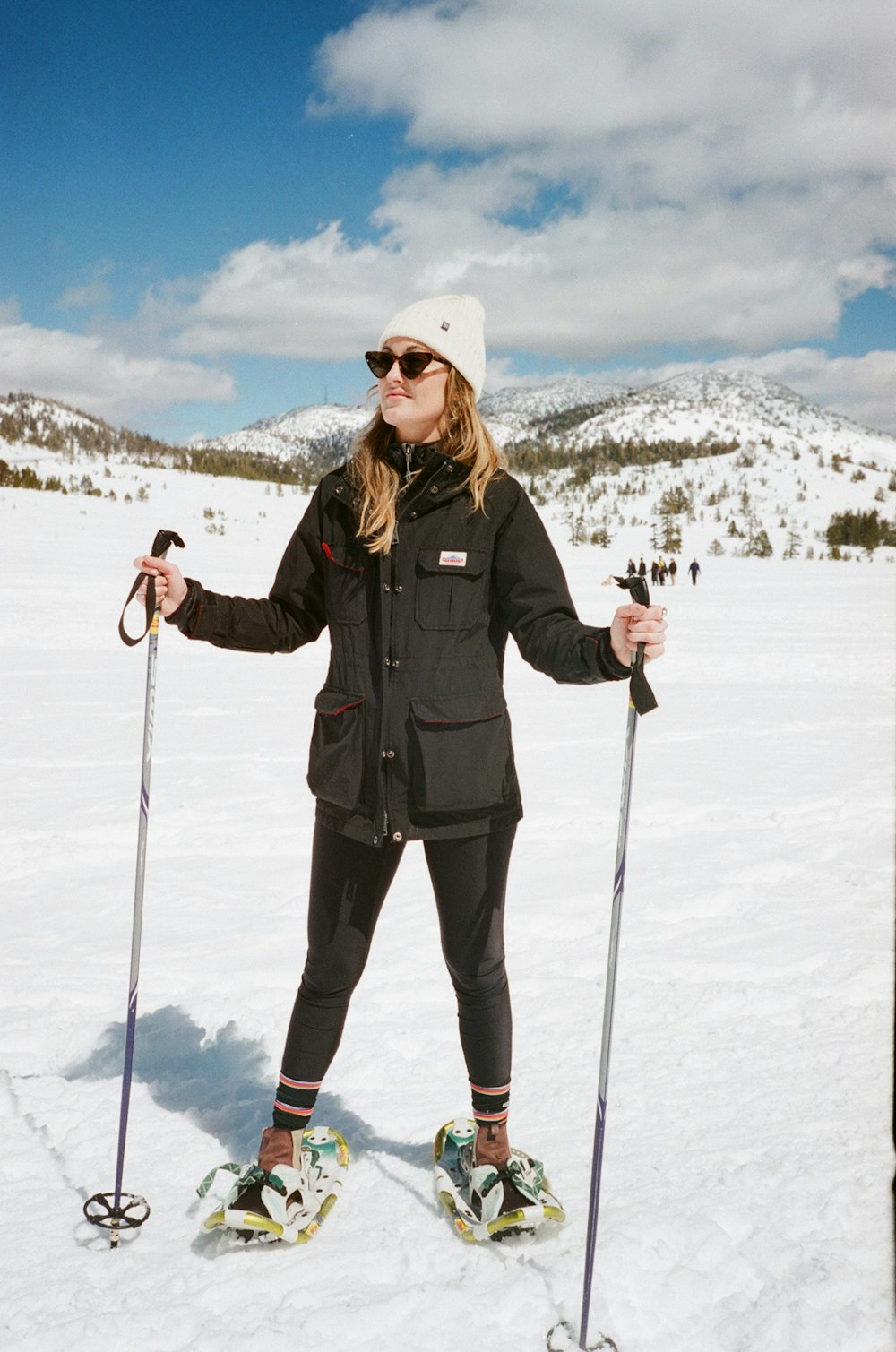 Mujer con chaqueta negra y pantalones negros con gafas blancas para la nieve de pie en el suelo cubierto de nieve