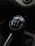A close-up view of a manual car gear shift knob displaying a five-speed transmission pattern, with numbers and letters in white on a black background. The interior of the vehicle has a dark tone, and the gearshift is centrally located.