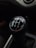 A close-up view of a manual car gear shift knob displaying a five-speed transmission pattern, with numbers and letters in white on a black background. The interior of the vehicle has a dark tone, and the gearshift is centrally located.