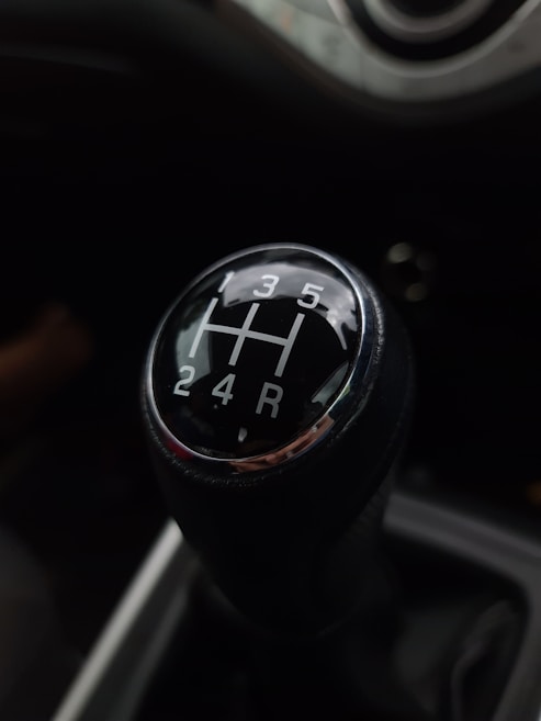 A close-up view of a manual car gear shift knob displaying a five-speed transmission pattern, with numbers and letters in white on a black background. The interior of the vehicle has a dark tone, and the gearshift is centrally located.