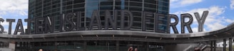 A large building entrance with modern architectural design features prominently, displaying the words 'Staten Island Ferry' in bold, metallic letters. The structure has a sleek, glass facade reflecting the sky and nearby structures.