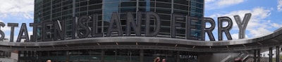 A large building entrance with modern architectural design features prominently, displaying the words 'Staten Island Ferry' in bold, metallic letters. The structure has a sleek, glass facade reflecting the sky and nearby structures.