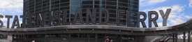 A large building entrance with modern architectural design features prominently, displaying the words 'Staten Island Ferry' in bold, metallic letters. The structure has a sleek, glass facade reflecting the sky and nearby structures.