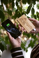 A person holds a smartphone showing a fully charged battery symbol connected to a wooden power bank. The power bank has a geometric logo and the word 'agefis' engraved on it. In the background, there are green leaves, suggesting a natural setting.