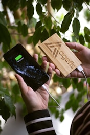 A person holds a smartphone showing a fully charged battery symbol connected to a wooden power bank. The power bank has a geometric logo and the word 'agefis' engraved on it. In the background, there are green leaves, suggesting a natural setting.