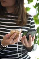 A person holding a smartphone in one hand and a wooden power bank with a white charging cable in the other. The person is wearing a striped shirt and appears to be standing near green foliage.
