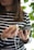A person holding a smartphone in one hand and a wooden power bank with a white charging cable in the other. The person is wearing a striped shirt and appears to be standing near green foliage.