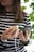A person holding a smartphone in one hand and a wooden power bank with a white charging cable in the other. The person is wearing a striped shirt and appears to be standing near green foliage.