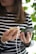 A person holding a smartphone in one hand and a wooden power bank with a white charging cable in the other. The person is wearing a striped shirt and appears to be standing near green foliage.