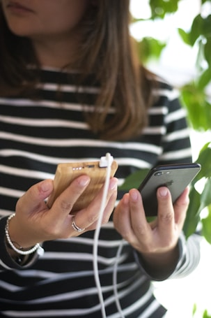 A person holding a smartphone in one hand and a wooden power bank with a white charging cable in the other. The person is wearing a striped shirt and appears to be standing near green foliage.