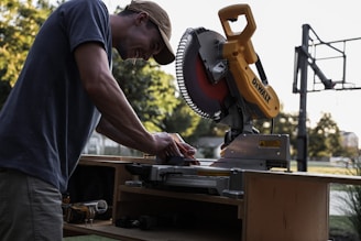 A person is working with a yellow and black miter saw on a wooden workbench outside, with trees and a basketball hoop visible in the background. The person is wearing a cap and appears focused on their task.