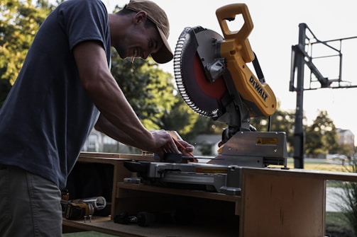 A person is working with a yellow and black miter saw on a wooden workbench outside, with trees and a basketball hoop visible in the background. The person is wearing a cap and appears focused on their task.