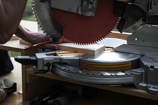 A circular saw is cutting a piece of wood with a person holding the wood in place. The saw blade is metallic, sharp, and partially visible with safety markings on it. The setting appears to be a workshop or construction site with tools around.