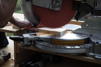 A circular saw is cutting a piece of wood with a person holding the wood in place. The saw blade is metallic, sharp, and partially visible with safety markings on it. The setting appears to be a workshop or construction site with tools around.