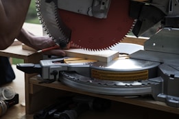 A circular saw is cutting a piece of wood with a person holding the wood in place. The saw blade is metallic, sharp, and partially visible with safety markings on it. The setting appears to be a workshop or construction site with tools around.