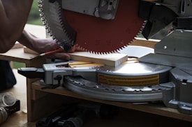 A circular saw is cutting a piece of wood with a person holding the wood in place. The saw blade is metallic, sharp, and partially visible with safety markings on it. The setting appears to be a workshop or construction site with tools around.
