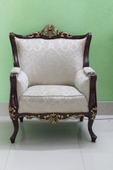 An elegant armchair with intricate wooden carvings and light-colored upholstered fabric featuring a subtle floral pattern. The armchair is placed on a glossy tiled floor against a light green wall.