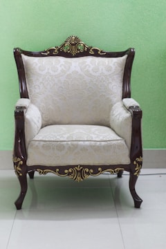 An elegant armchair with intricate wooden carvings and light-colored upholstered fabric featuring a subtle floral pattern. The armchair is placed on a glossy tiled floor against a light green wall.