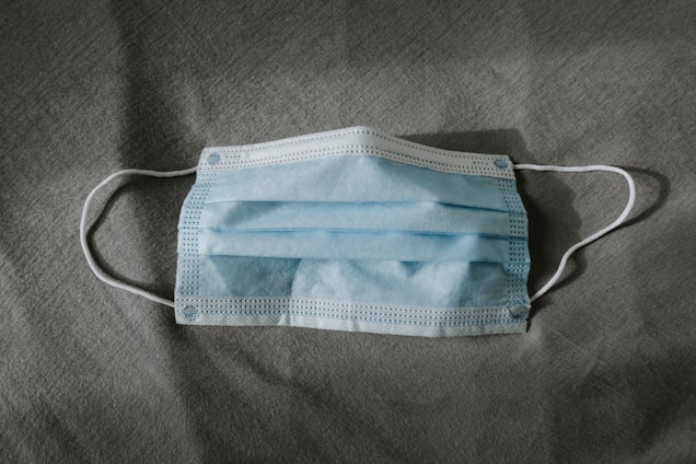 A blue disposable surgical mask is placed flat on a gray textured fabric surface. The mask has white ear loops and visible pleats.