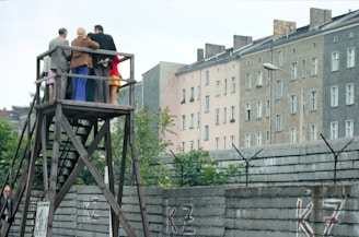 A group of people stands on a wooden platform, observing something in the distance. The structure is elevated, allowing them a clear view over a tall concrete wall topped with barbed wire. In the background, there are several multi-story residential buildings with numerous windows. The setting is urban and appears to be in a historical or tense context.