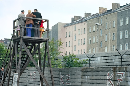A group of people stands on a wooden platform, observing something in the distance. The structure is elevated, allowing them a clear view over a tall concrete wall topped with barbed wire. In the background, there are several multi-story residential buildings with numerous windows. The setting is urban and appears to be in a historical or tense context.