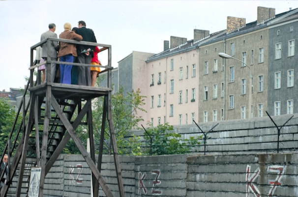 A group of people stands on a wooden platform, observing something in the distance. The structure is elevated, allowing them a clear view over a tall concrete wall topped with barbed wire. In the background, there are several multi-story residential buildings with numerous windows. The setting is urban and appears to be in a historical or tense context.