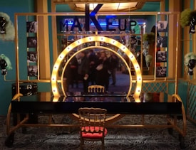An elaborate makeup station with a large illuminated circular mirror in the center, surrounded by a golden frame. A single ornate chair sits in front of the station. Photographs and decorative wigs are displayed on either side. Teal walls and elegant carpet flooring complement the luxurious setting.
