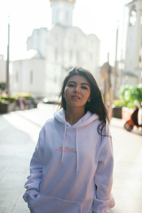 A person wearing a white hoodie stands confidently on a sunlit street. In the background, a large, bright building with architectural detail is softly blurred. The scene captures a casual yet vibrant urban setting.
