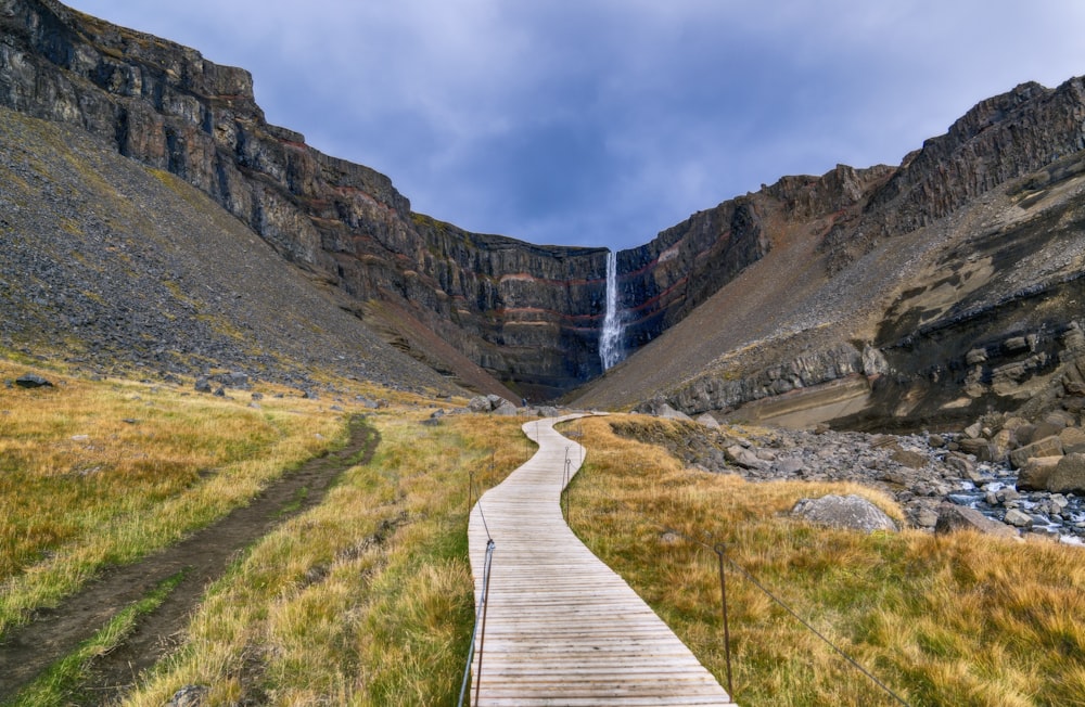 brown wooden pathway on green grass field near brown rocky mountain under white clouds during daytime