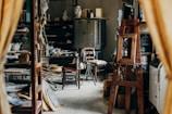 A cluttered workshop or studio filled with various objects, including wooden furniture, shelves with ceramics, art supplies, and a wooden easel. The lighting is soft, and the room has a cozy, lived-in feel.
