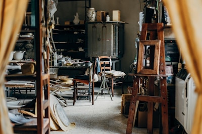 A cluttered workshop or studio filled with various objects, including wooden furniture, shelves with ceramics, art supplies, and a wooden easel. The lighting is soft, and the room has a cozy, lived-in feel.