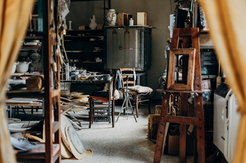A cluttered workshop or studio filled with various objects, including wooden furniture, shelves with ceramics, art supplies, and a wooden easel. The lighting is soft, and the room has a cozy, lived-in feel.