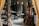 A cluttered workshop or studio filled with various objects, including wooden furniture, shelves with ceramics, art supplies, and a wooden easel. The lighting is soft, and the room has a cozy, lived-in feel.