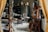 A cluttered workshop or studio filled with various objects, including wooden furniture, shelves with ceramics, art supplies, and a wooden easel. The lighting is soft, and the room has a cozy, lived-in feel.