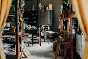 A cluttered workshop or studio filled with various objects, including wooden furniture, shelves with ceramics, art supplies, and a wooden easel. The lighting is soft, and the room has a cozy, lived-in feel.