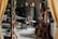 A cluttered workshop or studio filled with various objects, including wooden furniture, shelves with ceramics, art supplies, and a wooden easel. The lighting is soft, and the room has a cozy, lived-in feel.