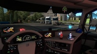 Inside the cabin of a truck, a detailed dashboard is visible with multiple controls and screens displaying maps and vehicle information. Ornamental figures, including a Santa Claus figure and a winter-themed character, are placed on the dashboard along with a cap. Outside the cabin, several parked trucks and greenery are visible, indicating a truck parking or rest area.