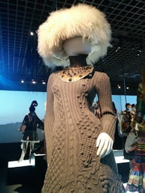 A mannequin wearing a detailed, textured brown knitted dress with a high neck and intricate patterns. The mannequin's head is covered with a large, fluffy white hat. The setting is a dimly lit gallery or exhibition space, with other mannequins in the background, each showcasing unique outfits.