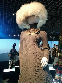 A mannequin wearing a detailed, textured brown knitted dress with a high neck and intricate patterns. The mannequin's head is covered with a large, fluffy white hat. The setting is a dimly lit gallery or exhibition space, with other mannequins in the background, each showcasing unique outfits.